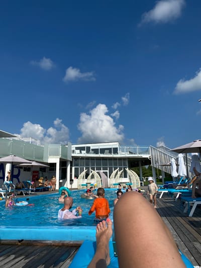 The image depicts a sunny day at an outdoor swimming pool complex. The pool is filled with blue water and several people are enjoying themselves. In the foreground, a person's legs and feet are visible, resting on a blue lounge chair. A wooden deck surrounds the pool.
In the pool, various individuals are engaged in different activities. Some are swimming, while others are using inflatable rings. A man in a white shirt is visible in the water, and a child in an orange shirt is also present. On the far left, a girl wearing goggles and a swimsuit is in the pool with an inflatable ring.
In the background, a modern building complex with multiple levels and large windows overlooks the pool area. Several white, dome-shaped structures are also visible, possibly cabanas or seating areas. Umbrellas provide shade around the pool and on the deck. The sky is a clear blue with scattered white clouds.
The location is identified as the city of Приамурский, Russia, suggesting this is a recreational facility within that region.