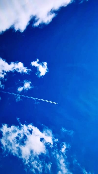 The image depicts a vibrant blue sky with scattered white clouds. A prominent detail is a contrail from an airplane, streaking diagonally across the frame from the bottom right towards the top. The contrail is long and thin, suggesting the plane is at a high altitude. The clouds vary in density and form; some are wispy and spread out, while others are more clustered and voluminous. The overall scene conveys a sense of open space and height, looking directly upwards. The bright blue of the sky indicates a clear day, likely during daylight hours. There are no visible people, text, or specific location cues within the image itself, beyond the general context of a sky scene.