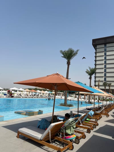The image depicts a resort swimming pool area in Ras Al-Khaimah, United Arab Emirates, on a clear, sunny day. In the foreground, several lounge chairs with cushions are set up on a tiled patio, some occupied by individuals relaxing. One chair has a baby stroller parked beside it. A large orange umbrella shades a portion of the patio.
The pool itself is a prominent feature, with clear blue water. A lifeguard station with a lifeguard in uniform is visible near the edge of the pool. Several rows of lounge chairs and umbrellas are lined up along the poolside. In the background, palm trees and a modern building with multiple floors are visible. A bird is captured in flight against the pale blue sky. The scene conveys a relaxed atmosphere typical of a resort vacation.