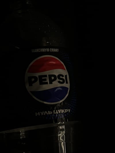 A dark, low-light image shows a bottle of Pepsi Max with Cyrillic text on the label. The bottle is partially illuminated, with reflections visible on its surface, suggesting a dark indoor environment. The Pepsi logo, a red, white, and blue circle with the word "PEPSI" in white, is clearly visible on the dark label. Above the logo, the text "МАКСИМУМ СМАКУ" (Maximum Taste) is present, and below it, "НУЛЬ ЦУКРУ" (Zero Sugar) is visible. The background is predominantly black, obscuring most of the surroundings. No people or specific location details within Podilsk, Ukraine, are discernible from the image.