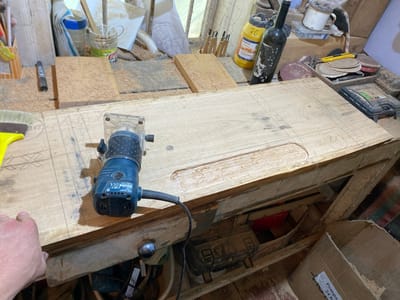 The image depicts a workshop environment in Negrești, Moldova, characterized by woodworking tools and materials. In the foreground, a person's left hand rests on a wooden workbench, indicating their involvement in a woodworking project. A blue and black Bosch router is positioned on the workbench, ready for use or recently used, with a visible power cord extending from it. The workbench itself is made of light-colored wood and is covered in wood shavings, suggesting recent or ongoing work. Several other objects are scattered around the workbench, including a yellow brush, a marker, a small block of wood, a bottle of yellow liquid labeled "30 Clet PVA-D2," a dark glass bottle, a white mug, various sanding discs, and a black toolbox. In the background, more tools and materials are visible, including jars, papers, and what appears to be a dismantled structure or shelving unit. The overall impression is of an active, albeit somewhat cluttered, workspace dedicated to woodworking.