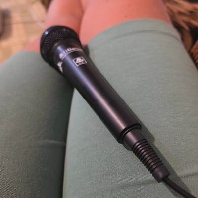 An overhead shot shows a black microphone resting on a person's legs, which are clad in light green leggings. The microphone's body is smooth and black, with a textured mesh at the top. A small, white logo with a circular design is visible on the microphone's surface. The cable extends from the base of the microphone and disappears out of frame. The background consists of blurred, warm-toned flooring and a textured brown surface, possibly furniture. The scene is indoors and appears to be a casual setting. The specific location within the city of Ravnogor, Bulgaria cannot be determined from the image.