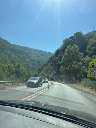 The image depicts a sunlit, mountainous landscape viewed from inside a vehicle traveling on a paved road. The road curves to the right, flanked by lush green hillsides covered in trees and rocks. Several vehicles, including a dark-colored car in the foreground and a white car further ahead, are visible on the road. Traffic cones and a guardrail are present along the side of the road. A yellow triangular warning sign with black symbols and text is visible on the right side of the road, indicating road conditions ahead. The sky is clear and blue, with the sun's glare creating a lens flare effect. Power lines are visible stretching across the sky in the upper right portion of the image. The scene appears to be in a rural or mountainous area, likely a road between towns or cities, given the absence of urban structures. Krupnik, Bulgaria is known for its mountainous terrain and historical significance, making it a plausible location for such a scene.