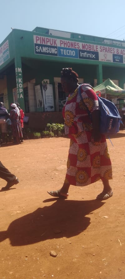 A daylight scene in Chemuche ward, Kenya, depicts an outdoor commercial area with a clear, bright blue sky. The foreground features reddish-brown earth, marked by pronounced shadows from direct sunlight.
The primary subject is a woman, viewed in profile, walking from right to left across the frame. She is attired in a full-length, red, white, and yellow patterned dress with a checkered design and circular sun-like motifs, complemented by dark sandals. A blue backpack, clearly marked with the "TECNO" logo in white text, is worn on her back. Her hair is styled in dark braids.
In the mid-ground, a single-story building with a green facade is visible. Its upper section displays a prominent sign reading "PINPUK PHONES, MOBILE SPARES & ACC..." directly above logos for "SAMSUNG," "TECNO," and "Infinix." A smaller sign for "ONFON MOBILE" is also present. Vertically on the left side of the building, a sign displays "M-PESA." Advertisements, including one for "SPARK 4G," are affixed to the building's windows. Several individuals are partially visible near the building's entrance, appearing stationary or engaged in conversation. Small plants in pots are arranged at the base of the building.
To the far right in the background, a green tent or canopy is partially visible. In the bottom left corner of the image, the leg and foot of another individual, wearing dark shoes, are partially visible, indicating additional pedestrian movement.