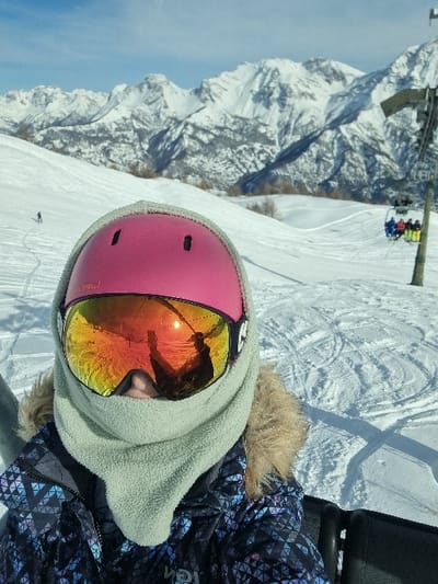 The image displays an individual on a snowy mountain slope in Cesana Torinese, Italy, positioned as if on a ski lift. The central subject, likely taking a selfie, wears a pink ski helmet with two visible black vents, mirrored orange and yellow ski goggles that reflect a person holding a device and a bright sun, a light grey fleece neck gaiter covering the lower face, and a blue, black, and white geometric-patterned ski jacket with a visible fur-lined collar. A black seat portion is visible to the subject's right. The midground shows a vast snow-covered terrain with numerous ski tracks. A lone skier is visible on the slope to the left. To the right, a dark ski lift pole supports a chairlift carrying at least three individuals. The background consists of a prominent range of snow-capped mountains under a clear blue sky, with sparse bare trees visible on the lower slopes.