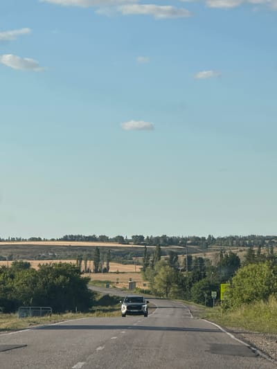 In Novotolucheyevo, Russia, a dark-colored SUV drives down a paved road on a clear, sunny day. The road curves to the right, leading towards a landscape of rolling hills covered in trees and fields of golden crops. The sky is a vibrant blue with a few scattered white clouds. Green foliage lines the sides of the road, and a small speed limit sign with the number "40" is visible on the right.