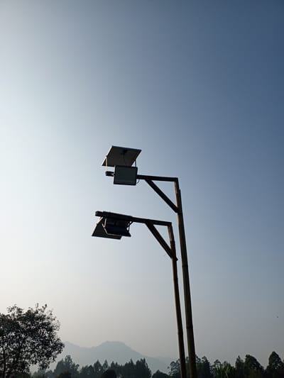 The image depicts a clear sky over a rural landscape, possibly in or near Bellanneoya, Sri Lanka. The sky transitions from a bright, pale blue near the horizon to a deeper blue at the top. In the foreground, a rustic lamppost made of what appears to be bamboo or weathered wood supports two solar-powered lights. Each light fixture is angled downward, with a rectangular solar panel positioned above it.
Below the lamppost, a dense line of trees and foliage forms the middle ground. These trees vary in shape and density, with some appearing as tall, slender pines and others as broader, leafy trees. Behind the treeline, rolling hills are visible, softened by a haze that suggests distance or atmospheric conditions. In the far distance, the hills recede into the pale blue of the sky. On the left side of the image, a large, dark tree with a full canopy stands out against the lighter sky. The overall scene conveys a sense of natural tranquility and the presence of renewable energy infrastructure in a rural setting. There are no people visible in the image.