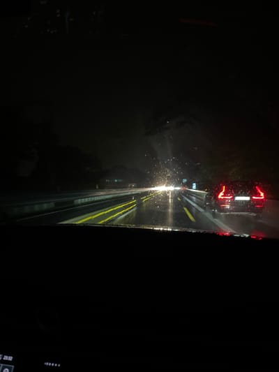 This image depicts a nighttime view from inside a vehicle on a wet road, possibly in or near Leverkusen, Germany. The road is illuminated by the vehicle's headlights and the taillights of a car ahead, a dark-colored SUV, which is partially visible on the right side of the frame. The road surface is reflective due to rain, with yellow lane markings visible. Distant lights and trees are visible on the left and right sides of the road, suggesting an urban or suburban environment. The windshield of the vehicle shows streaks of rain and light reflections. The overall atmosphere is dark and appears to be during a rainstorm.