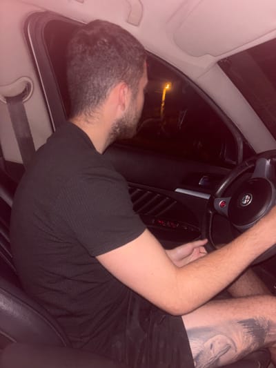 The image shows a man sitting in the driver's seat of a car at night. He is wearing a black t-shirt and dark shorts. A tattoo is visible on his left thigh. The car's interior is dimly lit, with the dashboard and steering wheel visible. Outside the car window, a street light and dark foliage can be seen, suggesting an urban or suburban setting in Каменар, Bulgaria. The man appears to be holding the steering wheel, possibly driving or preparing to drive. The scene is captured from a slightly elevated, side-view perspective.
