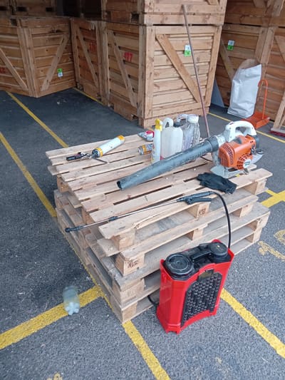 A scene is depicted outdoors in Biloziria, Ukraine, with a focus on gardening and cleaning equipment. Several large, wooden crates are stacked in the background. In the foreground, a collection of items is laid out on a stack of wooden pallets. These items include a leaf blower, a handheld sprayer, gloves, and several bottles and cans of liquid. A red backpack sprayer sits on the ground beside the pallets. The ground is paved with asphalt and marked with yellow lines. The lighting suggests it is daytime.