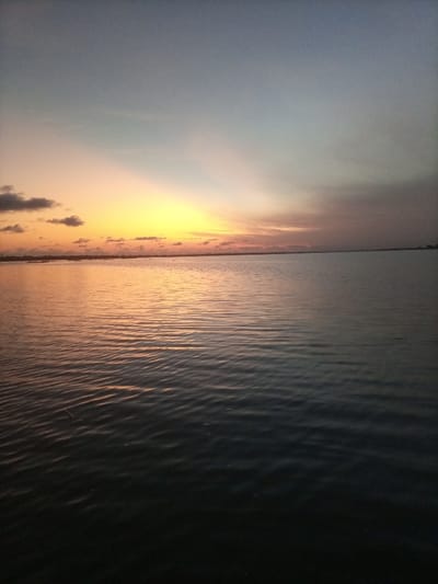 The scene depicts a wide body of water under a twilight sky, located in Akoko, Nigeria. The sky transitions from dark blue-gray at the top to a vibrant orange and yellow glow concentrated at the left horizon, which diffuses across the central horizon line into a softer pinkish-gray. Scattered dark cloud formations are present in the upper left quadrant, silhouetted against the brighter sky. The surface of the water displays numerous small, consistent ripples, reflecting the atmospheric colors; the foreground water is dark blue-gray, gradually becoming an orange-gold reflection closer to the horizon. A thin, indistinct dark line along the distant horizon suggests a landmass or shoreline. No individuals, animals, vessels, or distinct man-made objects are visible within the frame.