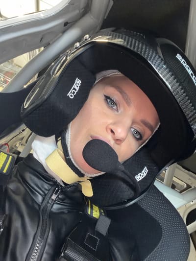 A female individual is depicted in a close-up, angled shot, seated within the cockpit of a vehicle in Meuspath, Germany. She presents with fair skin, blue eyes enhanced with dark eyeliner, and light-colored eyebrows. Her head is enclosed in a black carbon fiber racing helmet, which features "sparco" branding on the integrated black ear cups. A black microphone boom extends from the helmet, positioned in front of her mouth. A light-colored balaclava is visible beneath the helmet, and a yellow safety strap, consistent with a HANS device or racing harness, is visible around her neck. She wears a black zippered jacket over a white undergarment. The visible interior environment consists of a black racing seat with perforated sections on the right side and a silver-colored roll cage structure, including diagonal and horizontal bars, in the background.