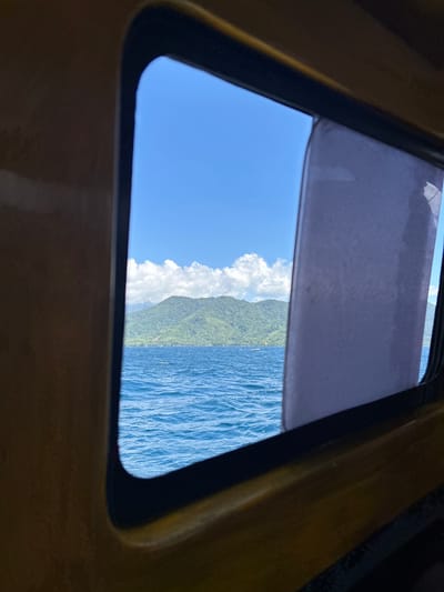 View through a boat window shows a body of water with ripples under a clear blue sky with scattered white clouds. In the distance, a lush green, mountainous island is visible, with a faint hint of buildings along its coastline. The window frame, made of dark material with a yellow accent, partially obstructs the view on the left and right sides. A translucent, light-colored shade is pulled down on the right side of the window. The overall scene suggests a journey at sea with a view of a tropical island, likely within the archipelago of the Tidore Islands, Indonesia.