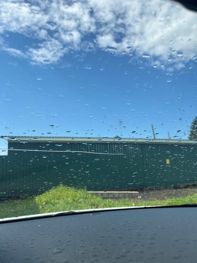 The image captures a view through a rain-streaked windshield. The sky above is a bright blue, dotted with fluffy white clouds. Below the sky, a long, dark green corrugated building with a lighter colored roof is visible. In front of the building, there is a patch of lush green grass. The foreground shows the textured surface of a car's dashboard, also speckled with raindrops. The scene appears to be an outdoor setting, possibly in or near the city of Mal'tsevo, Russia, given the context. There are no people or explicit actions depicted in the image.
