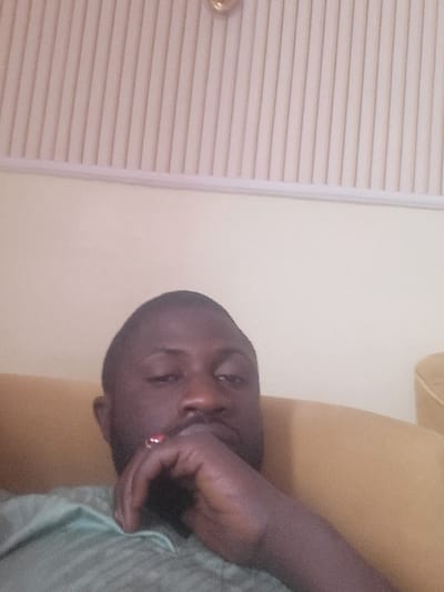 An adult male is depicted in an indoor setting, possibly in the city of Babawa, Nigeria. He possesses dark skin, short dark hair, and a beard, and wears a light green collared shirt. He is reclined on a light brown or tan upholstered sofa, with his right hand resting near his chin. A ring with a red stone is visible on his right ring finger. The background consists of a plain light cream-colored wall below a ceiling with vertical light-colored striped paneling. A small, circular, gold-colored fixture is partially visible at the top of the frame. The subject is looking directly towards the camera.