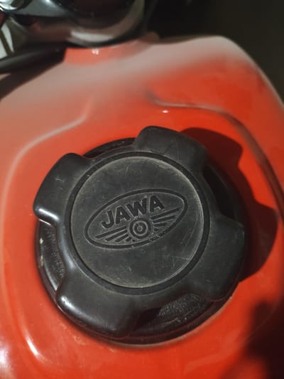 The image shows a close-up of a black fuel tank cap on a red motorcycle. The cap is circular with three protruding tabs for grip. The word "JAWA" is embossed on the cap, along with a winged emblem. The motorcycle's fuel tank is a reddish-orange color. The location of the image, Yerzhovo, Moldova, cannot be determined from the visual data provided.