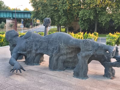 The image depicts a large, dark-colored sculpture of a reclining nude figure in the city of Haro, Spain. The sculpture is made of stone or a similar material and appears to be abstract, with rough textures and cave-like openings. The figure is lying on its back, with its head tilted upwards and an arm raised. One hand is outstretched, with fingers spread, resting on the paved ground. The sculpture is situated on a tiled walkway, with lush green foliage bordering the scene. In the background, a road curves away, lined with trees. A pedestrian overpass or bridge with a green metal structure is visible in the upper left portion of the image. The overall environment suggests an outdoor public space, possibly a park or promenade.