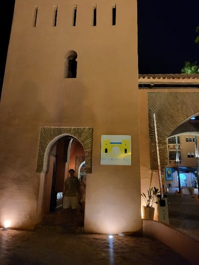 The image depicts a night scene at the entrance of a structure in Bouznika, Morocco, identified by a sign reading "PLAGE DE BOUZNIKA" (Beach of Bouznika). The main architectural feature is a light-colored, possibly ochre-toned, building or wall with traditional Moroccan design elements.

On the left, a prominent tower-like section features a crenellated top with five vertical slits and a keyhole-shaped decorative opening below them. At the base of this section, a pointed, ornate archway with intricate detailing serves as a pedestrian entrance. To the right of this archway, a wider, recessed archway is partially visible, featuring a textured ceiling and a terracotta-tiled roofline above it.

Two individuals are present within the left pedestrian archway. A male, wearing a light-colored short-sleeved shirt and shorts, stands centrally in the archway, facing the viewer. A second, partially obscured individual is positioned behind and to the right of the first, also within the archway.

A square white sign with a yellow border and a stylized building graphic, featuring the text "PLAGE DE BOUZNIKA," is affixed to the wall between the two archways. To the right of this sign, a white and red striped traffic barrier arm is in a raised position, connected to a dark grey barrier control unit. A small, green potted plant is situated near the base of the barrier unit.

The ground is paved with dark, patterned material and is illuminated by several ground-level spotlights that cast bright cones of light onto the architecture and ground. Artificial lighting is also present within the wider archway and on background structures. In the distant background to the right, additional buildings with illuminated windows and a blue poster are visible, along with the silhouettes of palm trees against a dark night sky.