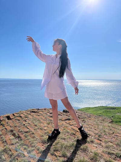 A vertical outdoor shot features a woman with dark hair wearing a white dress and black sandals. She stands on a brick and grass surface, seemingly near a body of water that stretches into the distance under a clear blue sky. The shot appears to be taken in Шуран, Russia, based on location. The sun's glare is visible in the upper left of the frame.