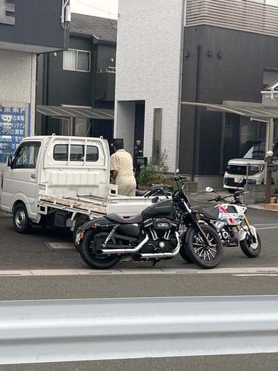 The image captures a street scene in Hanno, Japan, featuring a white utility truck parked on the left and two motorcycles positioned in front of it. A person in a light-colored uniform stands in the bed of the truck, facing away from the camera. The background includes buildings, a white van, and a grey guardrail. The scene is observed from across the street, with a portion of the guardrail in the foreground.
