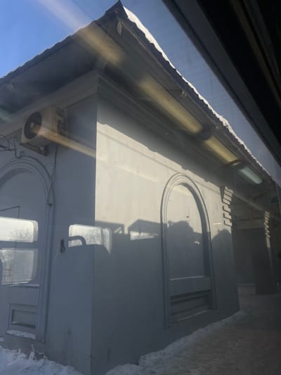 The image displays an exterior corner of a light-colored, single-story building located in Elektrougli, Russia, viewed through a reflective window. The building's facade is off-white or light grey, featuring a sloped roof with a slight overhang, partially covered by snow. A weathered outdoor air conditioning unit is mounted on the upper left wall, exhibiting rust streaks and discoloration. Below the air conditioner, an arched recess contains a rectangular window or panel. On the adjacent visible wall, another identical arched recess is present, appearing to be a decorative or sealed element. Two illuminated fluorescent tube lights are installed horizontally beneath the roof overhang. The scene is under strong sunlight from a clear blue sky, casting distinct shadows on the building's surfaces and the ground. The ground immediately surrounding the building's base is covered in a layer of white snow, with a cleared or dirty snow path visible in the foreground. No individuals or vehicles are observed within the frame.