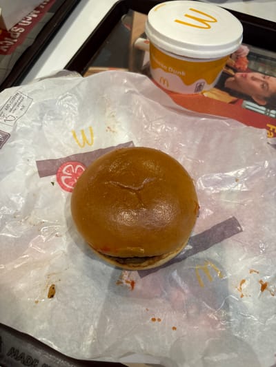 An overhead shot shows a McDonald's hamburger, still in its wrapper, and a container of McDonald's cheese sauce on a black tray. The wrapper is white and crumpled, with some sauce staining it. The hamburger is a standard McDonald's hamburger, and it appears to be slightly flattened. The container of cheese sauce is white with a yellow "M" logo. The tray is black. The image was likely taken indoors at a McDonald's restaurant, possibly in Bencongan Indah, Indonesia. A woman is also visible in the background.
