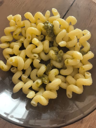 A top-down, close-up shot shows a clear glass bowl filled with bright yellow cavatappi pasta tossed with a vibrant green pesto sauce. The pasta is cooked al dente, with its distinctive corkscrew shape clearly visible. The pesto is generously applied, coating many of the pasta strands. The bowl rests on a wooden surface, likely a table, with a soft, diffused light illuminating the scene from above, suggesting it could be daytime. There are some reflections on the surface of the bowl, indicating a polished or glass-like material. The background is slightly blurred, emphasizing the pasta as the main subject. The overall impression is of a simple, appetizing dish, likely prepared for a meal.