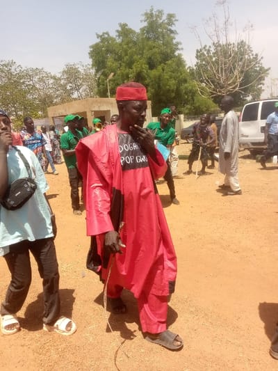 The scene depicts an outdoor gathering in Kagoma, Nigeria, under bright daylight with a clear sky. The ground is unpaved, appearing dry and reddish-brown.
In the foreground, a dark-skinned man is centrally positioned, facing slightly to his right. He wears a red traditional cap and a long, flowing red garment over a black undershirt. The black undershirt features white text that reads "DOGARI POP GWG". He has a short, dark beard and his right hand is raised, with fingers touching his chin. His left hand holds a thin, light-colored rope or cord. He wears dark sandals. To his immediate left, partially visible, is a younger individual wearing a light blue t-shirt, dark trousers, white sandals, and a black shoulder bag. This individual faces left, away from the central figure.
In the mid-ground, several individuals are present. A group of men, many wearing green shirts and green caps, are visible behind the central figure. One man within this green-clad group appears to be holding a shiny, brass wind instrument, possibly a trumpet or bugle, partially obscured. Further to the right of the central figure, another man in a light-colored (possibly off-white or light grey) traditional tunic and trousers stands, facing left. In the deeper mid-ground, another individual is seen holding a long stick or staff.
The background features a light-colored, possibly concrete or mud brick building with a flat roof, located to the left. Various green trees and sparse vegetation are scattered across the background, including a prominent tree with dense green foliage to the left and a bare, branchy tree to the right. A white passenger van is parked or moving on the far right side of the frame. Shadows are visible on the ground, indicating a high sun angle.