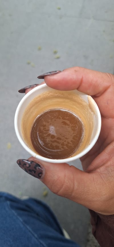 An overhead close-up shot captures a person's hand holding a small, white disposable cup filled with coffee. The hand exhibits a medium skin tone, and the fingernails are almond-shaped, adorned with a distinctive dark brown and black leopard print design. The cup contains a rich, dark brown liquid, indicative of espresso or strong coffee, with a prominent layer of light brown crema and small bubbles visible on its surface. The liquid occupies roughly half to two-thirds of the cup's volume.
The background is softly blurred, presenting a light gray, textured surface, possibly concrete or pavement, scattered with indistinct yellowish-green specks. This suggests an outdoor or casual urban setting, consistent with common scenes in Santa Cruz de Tenerife, Spain. A small section of blue fabric, likely denim jeans, is visible in the lower right corner, indicating the person may be seated. The image portrays a simple, casual moment, possibly a coffee break. No text is visible within the frame.
