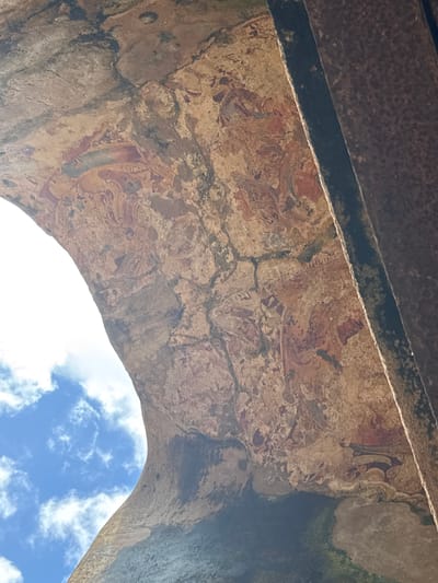 The image shows a section of the Gal Vihara, a rock temple complex in Polonnaruwa, Sri Lanka, from a low angle. The view is through a stone archway, with the sky visible beyond. The walls of the archway are weathered and cracked, revealing remnants of ancient paintings. The paintings, which feature human figures and decorative patterns, are in a faded state. The top of the archway is lined with a dark brown material. The sky is blue with scattered white clouds.