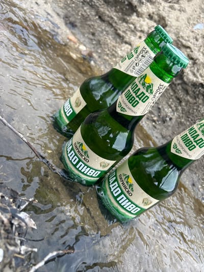 Three 330ml green glass bottles of 'Пиринско Младо Пиво' (Pirinjsko Mlado Pivo) with green caps are partially submerged in shallow, clear, flowing water. The bottles are positioned diagonally across the frame, from the lower left to the upper right. Their labels, which include a mountain emblem and text in Cyrillic, indicate the product name and state "ПАСТЕРИЗИРАНО" (Pasteurized). Adjacent to the water on the right, a textured bank of light brown or grey sand/dirt is visible. Minor organic debris, including a thin branch, is present in the foreground water. No individuals or specific man-made structures are discernible. The scene depicts an outdoor natural environment. While the requested location is Любен, Bulgaria, no identifiable landmarks or geographic features specific to that city are present within the visual content of the image to confirm this.