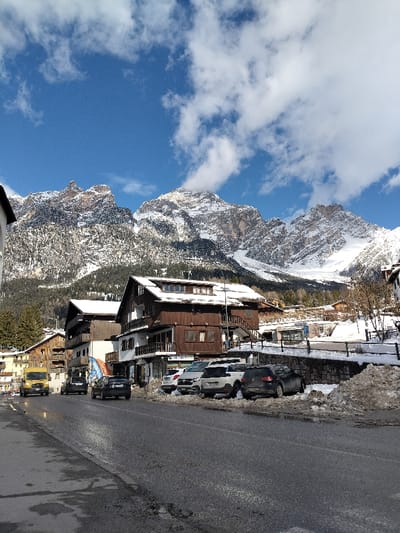 The image displays a winter or early spring street scene in Serdes, Italy. The foreground features a two-lane asphalt road, which appears damp with reflections, indicating moisture. Along the left side of the road, a yellow commercial van and a dark-colored sedan are in motion, proceeding away from the viewer. On the right side of the road, a row of approximately five parked vehicles, including several SUVs in white, dark gray, and black, are positioned perpendicularly to the curb. These vehicles are partially surrounded by accumulations of melting, soiled snow.

Dominating the mid-ground on the right are multiple multi-story buildings constructed in an alpine architectural style. The most prominent structure is a brown wooden building with white-railed balconies on several levels and a gabled roof heavily laden with snow, featuring multiple dormer windows. To its left, a white building with darker balconies is also visible, and all building roofs within the frame are covered in snow.

The background is defined by large, rugged mountains, entirely covered in snow and ice, with visible rock formations and patches of evergreen trees on their lower slopes. The sky above is a clear blue, adorned with scattered white cumulus clouds. No individuals are visible in the scene. The overall lighting suggests a sunny day.