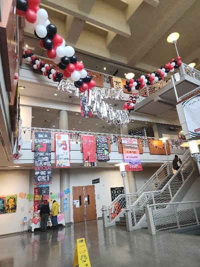 The image displays the multi-level atrium of the Seattle University Student Center in Seattle, Washington, USA. A long garland of red, white, and black balloons is strung across the upper sections of the atrium, with silver foil balloon letters spelling "RED HAWK" or a similar phrase beneath them. Various rectangular banners hang from the second-floor railing, displaying text such as "BE A RED HAWK", "HOMECOMING WEEK", and "Sustainable Student...". On the ground floor, two white-clothed tables are set up to the left. Two individuals are present at these tables: one wearing a dark jacket and a red beanie, and another in a yellow jacket, both focused on the table's contents. To the far left, a colorful art piece featuring two stylized faces is mounted on the wall. A yellow "WET FLOOR" caution sign is visible on the polished light-colored floor in the foreground. On the right, a multi-flight staircase with white metal railings leads to upper levels. A single individual in dark clothing is observed ascending this staircase. Double wooden doors are located behind the tables. The ceiling features a coffered design with visible beams.