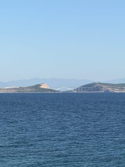 The image shows the Krk Bridge, a concrete arch bridge connecting the Croatian island of Krk to the mainland near the city of Kostrena. The left side of the image depicts a clear blue sky, while the right side captures the Adriatic Sea. The bridge's structure, including its arches and supporting pillars, is visible against the backdrop of the Krk island's coastline.