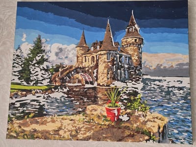 This is a painting of a castle on a rocky island in a body of water, possibly a lake or sea. The castle is made of stone and has several towers with conical roofs. There is a water wheel on the side of the castle. The water is depicted with choppy, dark blue and white brushstrokes, suggesting movement. On the shore in the foreground, there is a red pot with a green plant. The sky is a deep blue with streaks of lighter blue and some white clouds. The overall impression is of a dramatic and somewhat moody landscape, possibly at dusk or under stormy skies, given the dark sky and the choppy water. The style of painting appears to be a paint-by-numbers or similar hobbyist art, characterized by distinct areas of color and visible brushstrokes. There is no visible text.