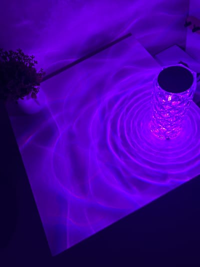 A detailed interior scene, photographed in Dmitriyevskaya, Russia, displays a light-colored rectangular surface, likely a table or desk, predominantly illuminated by a faceted, crystalline lamp. The lamp, positioned on the right side of the surface, emits vibrant purple light that refracts and projects swirling, concentric, and wave-like patterns across the surface and onto the adjacent background wall. A small potted plant with dark foliage is visible in the upper left corner, resting on the same surface. The entire visible environment is saturated with a uniform purple hue, indicating low ambient light with the lamp as the primary illumination source. No individuals are present.