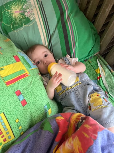 An infant is observed lying supine in a wooden crib within a residence in Романята, Russia. The infant, dressed in light-wash denim overalls featuring yellow bunny embroidered patches and "cute bunnies" text, holds a clear baby bottle with a yellow cap and nipple containing a white liquid. The crib is furnished with multiple textile items: a green pillow or blanket with a pattern of yellow, red, and blue geometric shapes on the left, a green sheet with a large yellow and white floral design and vertical green, black, and pink stripes on the top and upper right, and a folded multicolored blanket with purple, red, and yellow abstract patterns in the lower right foreground. Wooden crib slats are visible on the right side of the image. The infant's eyes are open and directed towards the viewer.
