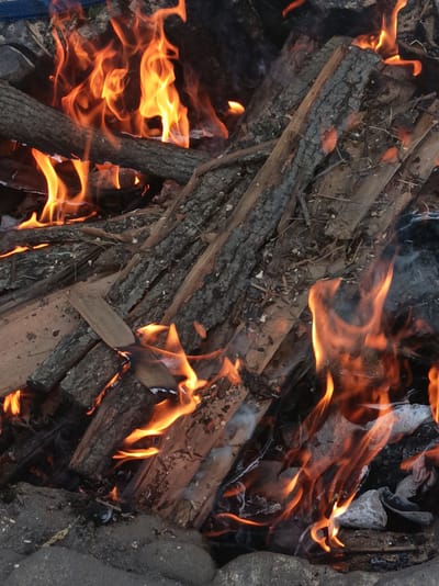 The image displays a close-up, overhead view of a campfire. The fire is burning brightly, with orange and yellow flames licking at several pieces of wood. The wood appears to be a mixture of larger logs with bark still attached and smaller, split pieces of firewood. Some of the wood is charred, indicating it has been burning for some time. The ground surrounding the fire is composed of dirt and small stones, consistent with an outdoor setting. There are no people visible in the image. The scene is likely a rural outdoor area, possibly a campsite or backyard, within the Шарпілаўскі сельскі Савет in Belarus.