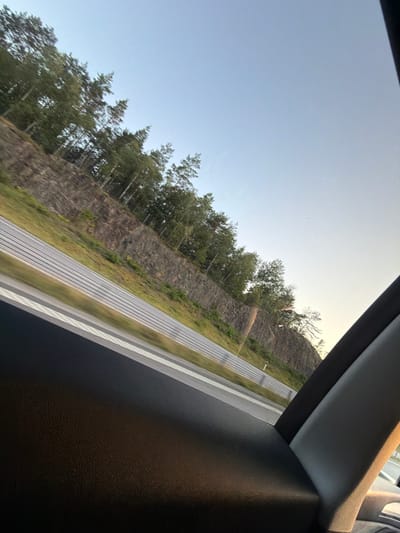 The image shows a view from inside a vehicle looking out at a highway embankment. The embankment is covered with grass and trees, and a rock face is visible on the upper portion. The highway itself has multiple lanes and is bordered by a guardrail. The sky is clear and blue. The image appears to have been taken on a sunny day, likely in the summer. The location is given as the E6 highway near Munkedal, Sweden.