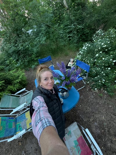 In Ozerki (Krasnodolinskaya), Russia, a woman with blonde hair pulled into a bun is taking a selfie outdoors. She is wearing a pink and white plaid shirt and a black puffer vest. She is standing near a blue table set with a bouquet of purple lupines in a vase, a silver kettle, and two white cups. Several folding chairs are arranged around the table. The scene is set in a wooded area with dense green foliage and white flowering bushes.