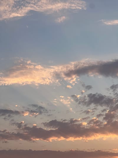 This is a vertical, medium-shot photograph of a sky filled with clouds at sunset. The sky transitions from a pale blue at the top to a soft orange and pink hue near the horizon, which is not visible in the frame. Wispy, thin clouds are scattered across the upper portion of the image, tinged with pink and white from the setting sun. Below these, larger, more textured clouds are present, some appearing dark gray and others illuminated with warm orange and yellow light. Rays of sunlight emanate from behind some of the clouds, creating a dramatic effect. The clouds are varied in shape and density, with some appearing fluffy and others more layered and streaky. There are no people, structures, or discernable landmarks visible in this image, as it solely captures the atmospheric conditions of the sky. The scene is devoid of any specific location details within Rotmanka, Poland, and offers no information about actions or interactions.