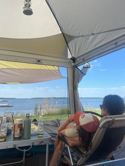 The image shows a campsite by the sea in the city of Am Salzhaff, Germany. In the foreground, a table is set up with two bottles of beer, a jar of coffee, a green water bottle, and other items. Two people are seated in chairs, facing away from the camera, looking out at the water. One person is wearing a red and white baseball cap. The background shows a body of water with boats and a distant shoreline. The sky is blue with some clouds. The overall scene depicts a relaxed outdoor setting.