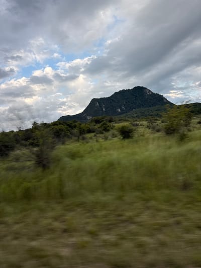 The image shows a mountain landscape with a cloudy sky. The mountain is covered with dense green trees and vegetation. A vast expanse of green grass is visible in the foreground, with the motion blur indicating movement, possibly from a vehicle. The sky is filled with dramatic, heavy clouds, with some patches of blue sky peeking through. The lighting suggests it could be late afternoon or early evening, with the sun low in the sky. The overall impression is one of a wild, natural landscape, possibly experienced during a journey. No people, text, or specific objects are clearly discernible. The scene evokes a sense of natural beauty and perhaps a feeling of being on the move through a picturesque, remote area.