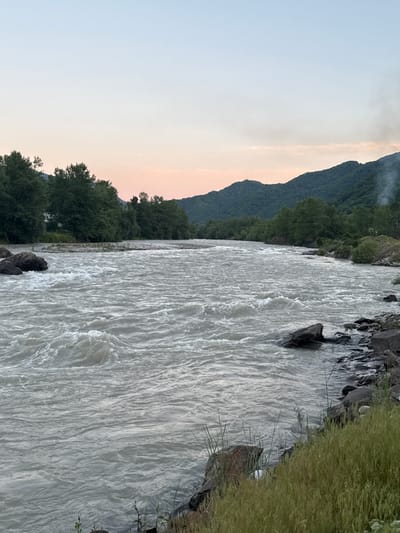 This is a landscape photograph taken in Kamenomostsky, Russia. The image depicts a wide, fast-flowing river with turbulent, white water. The riverbanks are lined with dark, wet rocks and lush green grass. In the background, a dense forest covers the slopes of a large mountain range. The sky above is a pale, hazy gradient, transitioning from a soft pinkish hue near the horizon to a light blue at the top, suggesting either sunrise or sunset. Some light mist or clouds are visible near the mountain peaks. The overall scene conveys a sense of wild, natural beauty and tranquility, characteristic of a mountainous, riverside setting. There is no visible text in the image.