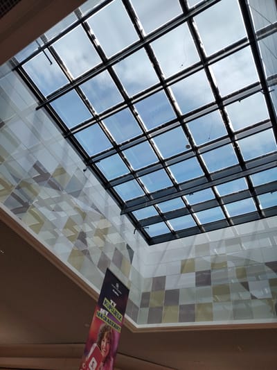 This media file shows an interior view of a modern building with a large glass-paneled ceiling, revealing a bright blue sky with scattered clouds, suggesting a daytime setting. The ceiling is framed by a dark grid structure. The interior walls are decorated with a mosaic of square tiles in varying shades of white, grey, and light green, creating a geometric pattern.
Hanging from the ceiling is a banner with text that appears to be in Polish. Partially visible text on the banner includes "GALERIA WOŁOMIN" and what seems to be a slogan or promotional message, potentially related to a shopping or retail environment. A portion of a person's face and shoulder is visible at the bottom of the banner, smiling and holding a red object.
The overall scene conveys a sense of contemporary architecture and a well-lit indoor space. The presence of the banner and the nature of the tiled walls suggest a public or commercial location, such as a shopping mall or a large public building in Wołomin, Poland. The lighting and clear sky indicate pleasant weather conditions.