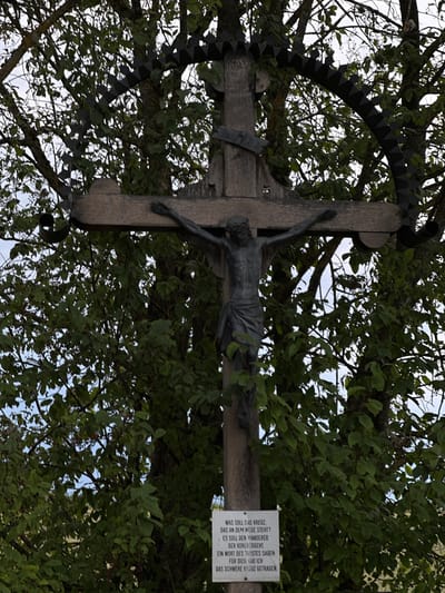 The image shows a wooden crucifix with a metal sculpture of Jesus Christ, set against a backdrop of lush green foliage and trees. The crucifix is mounted on a rough wooden pole. A black, ornate metal structure with pointed elements encircles the upper part of the crucifix. At the base of the crucifix, a white sign with black text is attached to the pole. The text on the sign appears to be a religious inscription, written in German. The environment suggests an outdoor setting, possibly in a rural area or a park, with the sky visible through the leaves. The sign is located in Rudelzhausen, Germany.