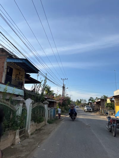 This is an eye-level, medium shot of a street in Alurcucur, Indonesia. The sky is a clear blue with thin white clouds. On the left side of the street, there is a two-story brick building with a yellow facade and a blue awning. A fence with green foliage runs along the front of the property. The street is paved and there are several power lines overhead. A motorcycle with two riders is moving away from the camera down the street. A white SUV is driving in the distance. There are also some wooden structures on the right side of the street. A woman in a white shirt and patterned pants is standing on the right side of the street, further down. A motorbike with a sidecar is parked on the right side of the road. The overall scene depicts a typical street in a rural Indonesian town.