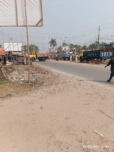 This image depicts a roadside scene in Kaliganj, Bangladesh, captured on March 12, 2025, at 11:01 AM.
The foreground on the left features a dirt and gravel shoulder heavily littered with various types of trash, including plastic bags and debris, alongside sparse dry vegetation. A large white rectangular signboard, supported by two bamboo poles, is partially visible in the upper-left, extending out of frame.
A paved asphalt road runs through the midground, curving slightly from left to right. Several vehicles are present on or beside the road: a yellow truck with a colorful blue cargo bed featuring Bengali text is positioned on the left side of the road; another yellow truck is partially visible further down; and a dark blue truck with a wooden cargo bed is parked on the right side of the road, with what appears to be logs or timber piled next to it. A motorcycle is visible near the center of the road, and a bicycle-rickshaw or similar vehicle is also present.
In terms of people, one individual, dressed in dark pants and shoes, is walking from right to left in the lower-right foreground. Other indistinct figures are visible near the trucks and roadside structures in the midground-left.
The background displays a typical urban or semi-urban landscape with numerous utility poles and crisscrossing power lines. Various commercial billboards and signs, some featuring images of individuals, line the far side of the road. Trees and sparse foliage are visible beyond the structures. The sky is clear, indicating a bright, possibly sunny day.