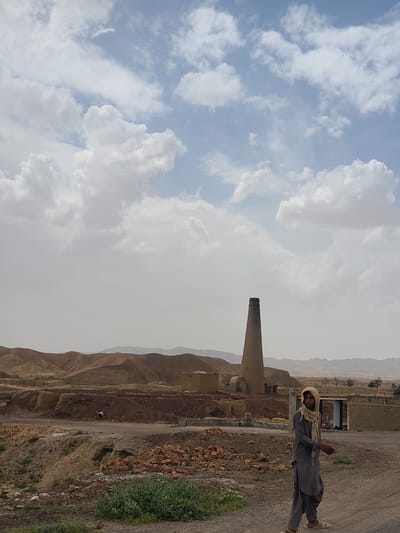 A vertical shot depicts a man standing to the right of a large chimney structure at a brick-making factory in the city of Unknown, Pakistan. The man is dressed in gray and has a light scarf around his head and neck. He faces the viewer, and the background consists of an arid landscape with a smokestack. The sky overhead features scattered clouds.