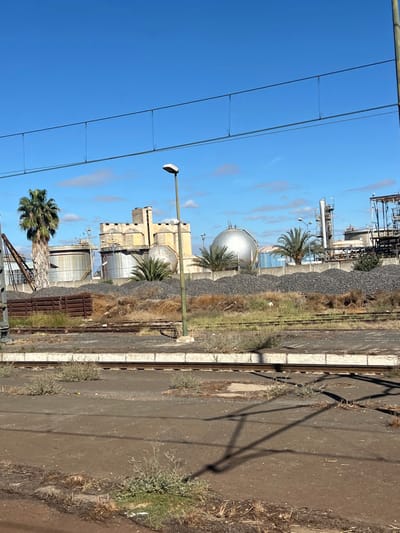 This image depicts an industrial landscape adjacent to a railway line in Sidi Kacem, Morocco, under a clear blue sky with scattered white clouds.

In the foreground, multiple railway tracks with concrete ties are visible, appearing unused due to sparse weeds and dry vegetation growing between and around them. A distinct pattern of shadows, cast by an unseen overhead structure or wires, stretches across the tracks and ground.

The midground features a low, concrete railway platform running parallel to the tracks. A slender, olive-green utility pole with a light fixture at its apex stands on this platform. Beyond the platform, the ground rises slightly, covered in dry vegetation and several large piles of dark, granular material, possibly coal or aggregate.

The background is dominated by an industrial complex. Prominent features include several large, metallic cylindrical storage tanks and two spherical storage tanks, one notably larger and reflective. Behind these tanks, a multi-story, light-colored industrial building with crenellated architectural details, resembling a castle or fort, stands centrally. To the right, additional industrial structures, including towers, piping, and scaffolding, are visible. Several palm trees of varying heights are interspersed throughout the industrial area. No individuals or active movements are discernible within the scene. The lighting suggests bright daylight.